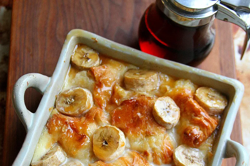 Ingredients for banana croissant casserole including torn croissants, sliced bananas, eggs, milk, cream, and spices arranged on a marble countertop